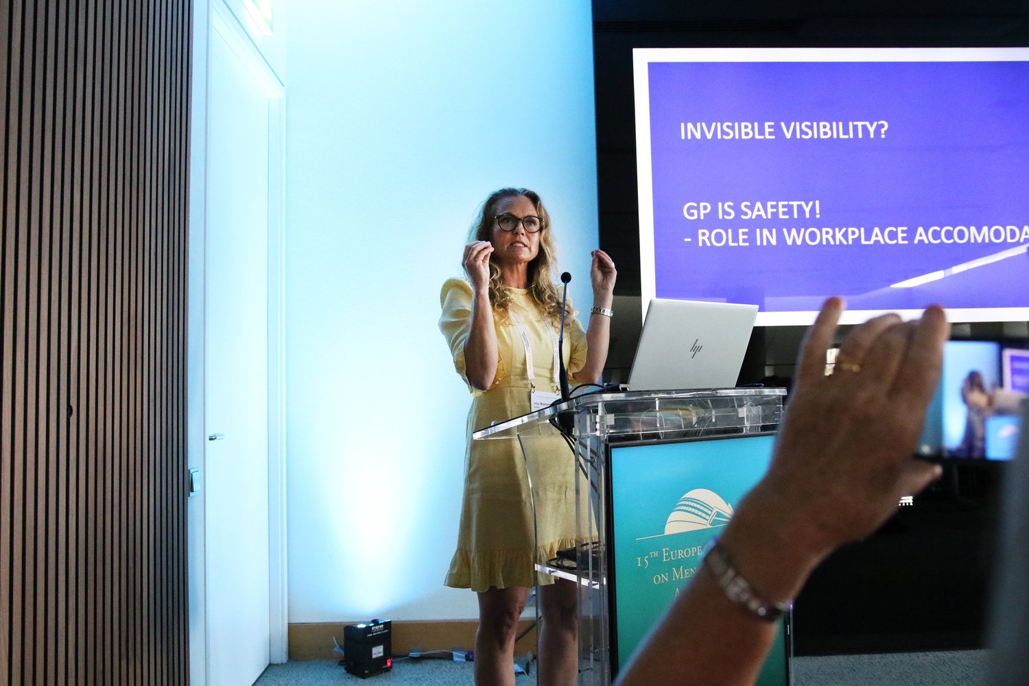 kvinne gestikulerer mens hun prater til en forsamling, mens noen tar bilde av henne med mobiltelefonen sin