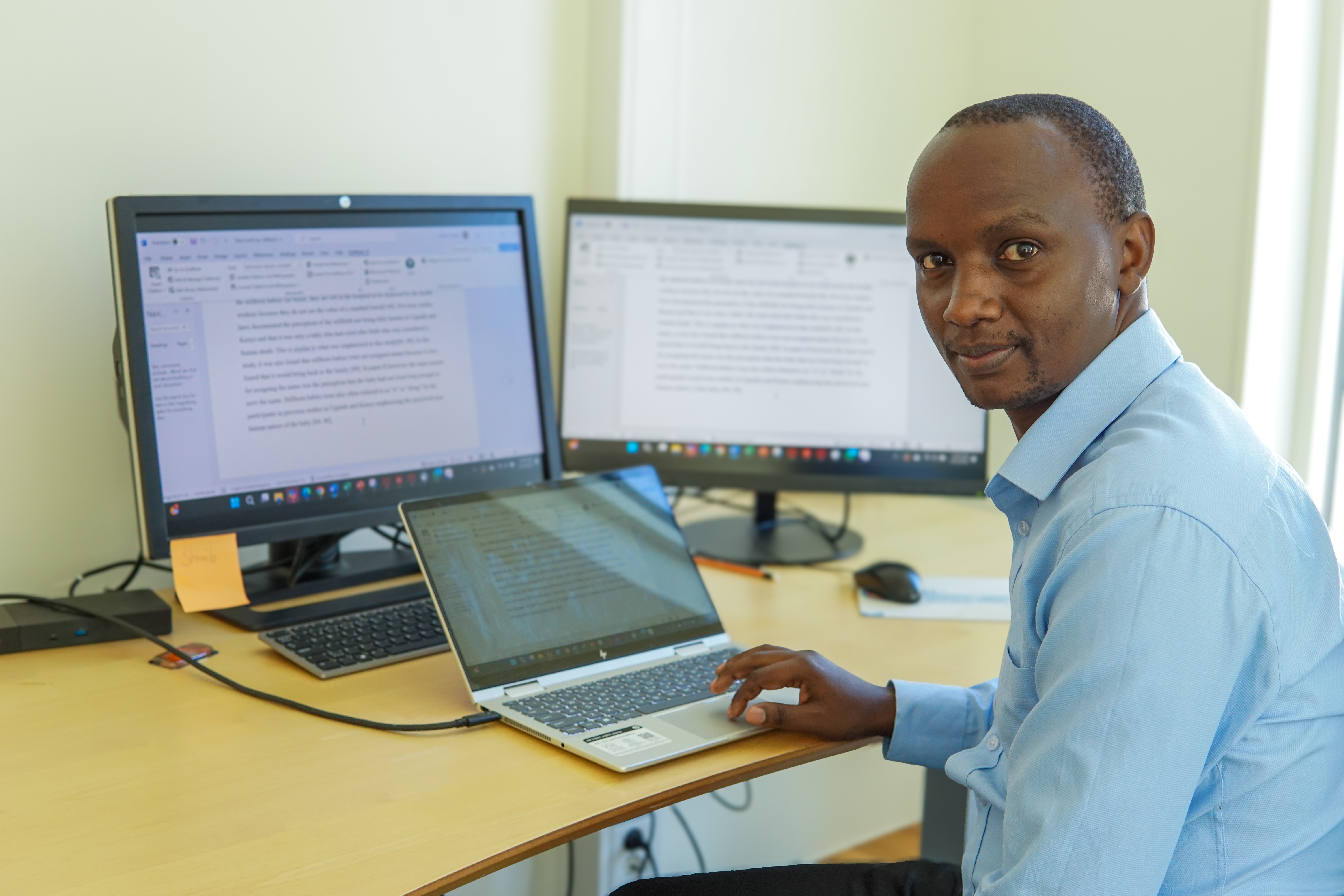 Bilde av stipendiat Martin Chebet ved arbeidsbordet sitt. Skjermene er åpne og synlige. Chebet ser mot kamera 