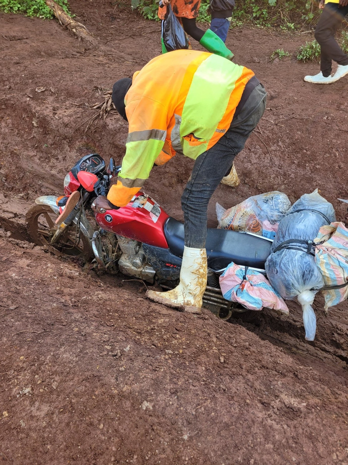 Photograph showing a person wearing a bright orange and yellow jacket and white boots struggling to lift a red motorcycle stuck in a deep muddy trench. The motorcycle is loaded with multiple large bags tied to the back, and other people with boots are visible in the background on rough, muddy terrain.