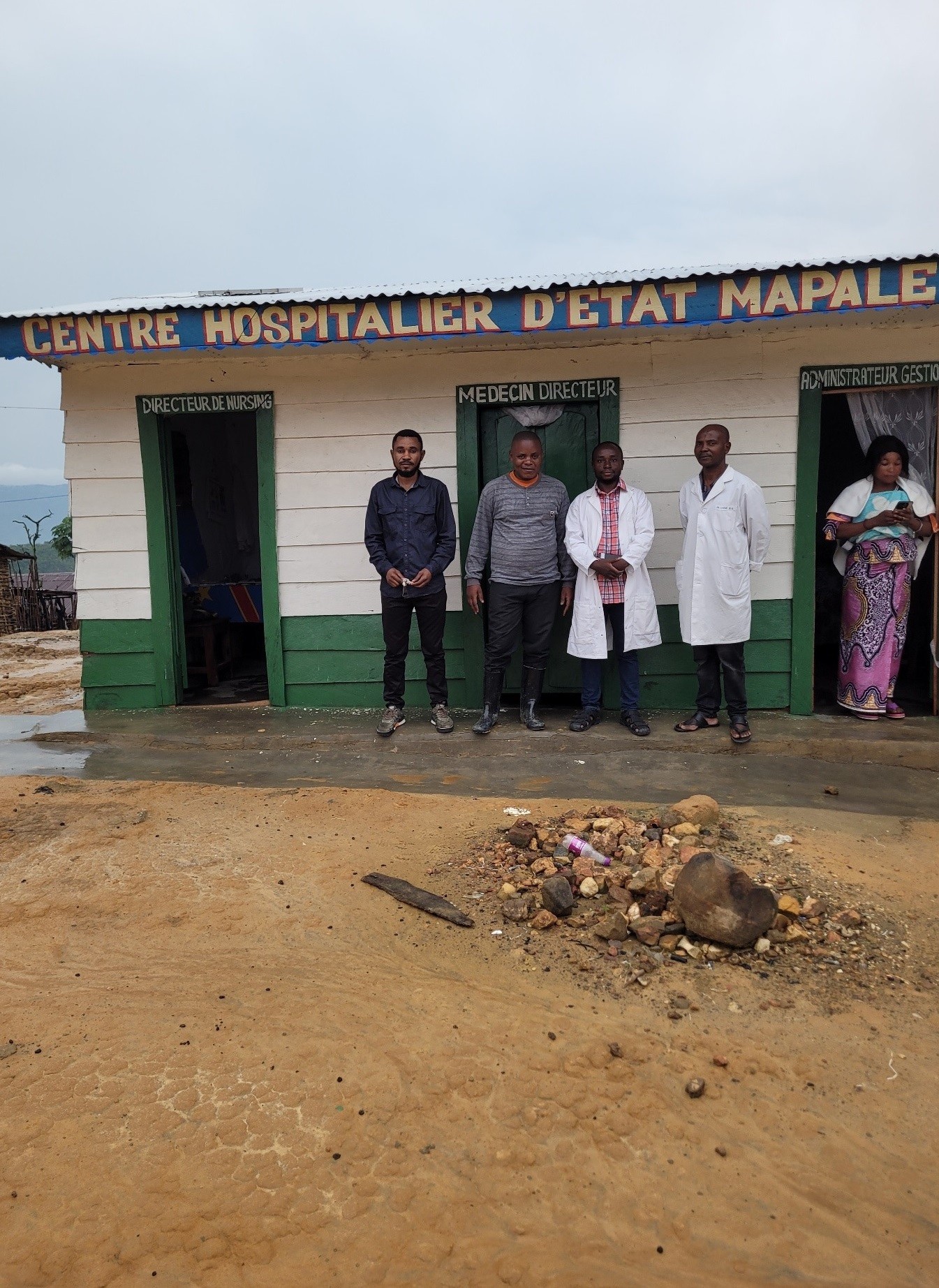 Photograph of five people standing outside a small rural hospital building." The building has three green doors and the ground in front shows a patch of disturbed earth with scattered debris.