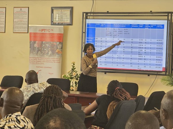 Woman standing, holding microphone and pointing to meeting screen in front of people attending meeting.