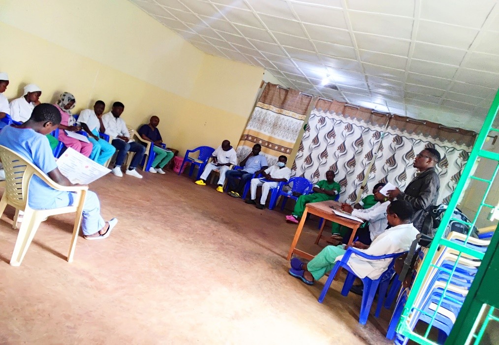 Photograph of a group meeting in a large room with people seated in a semi-circle on plastic chairs, some wearing medical scrubs and others casual clothing. A person stands near a table addressing the group, indicating a discussion or training session in a healthcare or community setting.