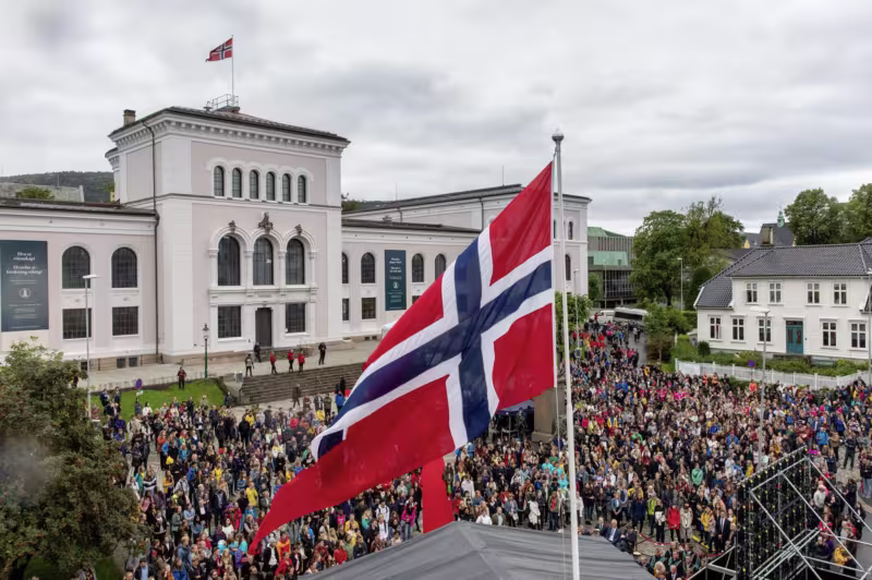 Norsk flagg i forgrunn, med nye studenter i bakgrunn foran Universitetsmuseet i Bergen på en grå overskyet dag, i forbindelse med åpning av nytt studieår.