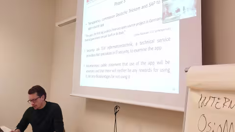 En mann i svarte briller og svart genser foran et lerret som viser en presentasjon