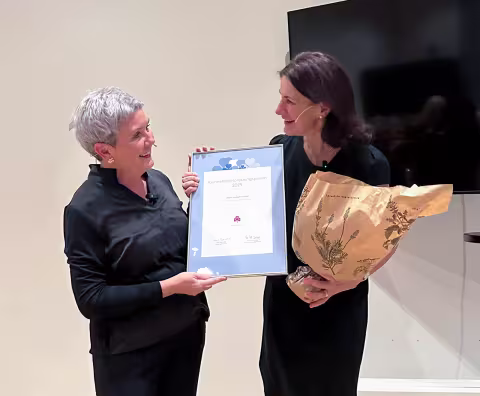 To kvinner på scenen, der den ene gir et diplom og blomster til den andre.