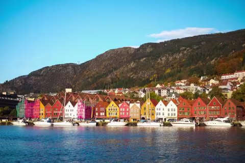 Bilde av bryggen i Bergen fra tatt fra avstand. Man ser også deler av Vågen med båter som ligger til kai og fjellet bak bryggen. 