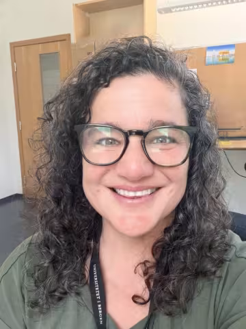 Profile picture, woman with dark curly hair and glasses, smiling. Office environment in the background.