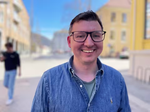 Mann med brunt hår og firkantete briller smiler inn i kamera