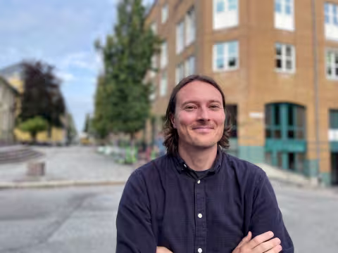 Mann med halvlangt blondt hår smiler inn i kamera. Han står utenfor SV-fakultetet