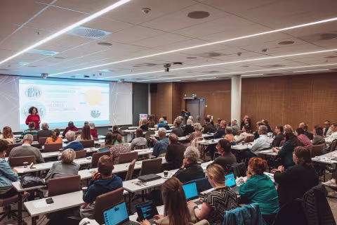 Group of people watching a woman presenting something on a screen.
