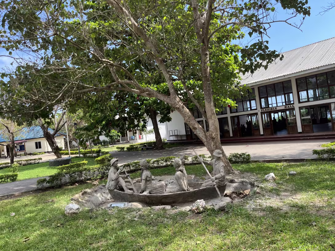 A tree, and a scultupture showing four people in a boat, outside a building. The building hosts the Bagamoyo Institute of Arts and Culture (TaSUBa) in Tanzania