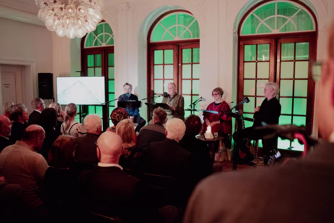 Publikum som ser på fire menneske som les frå eit stykke, ein skjerm til venstre for dei