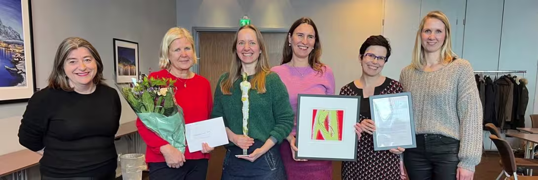 A group of colleagues gathered for a celebration. One person is holding a bouquet of flowers and an envelope, while another holds a trophy. Two others are displaying framed artwork and a certificate. They are standing in a meeting room with tables and chairs, and a scenic photograph is visible on the wall in the background