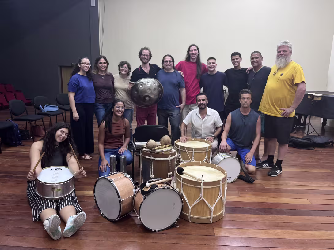 A group of people pose together in a rehearsal room, surrounded by various drums and percussion instruments arranged on the floor in front of them.
