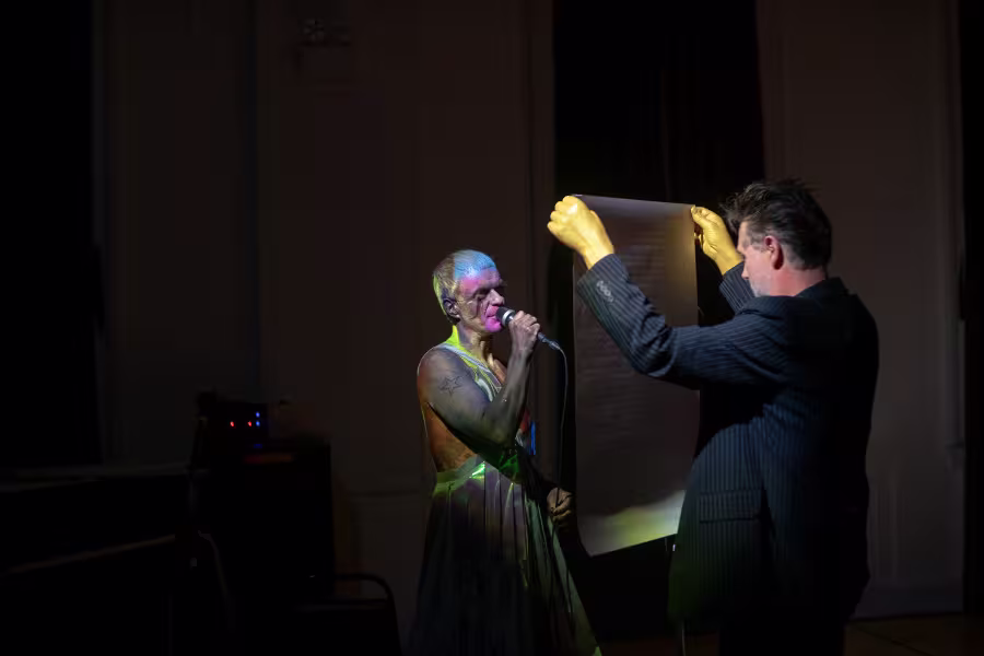 Two performers on a dark stage, one speaking into a microphone while another holds up a large reflective panel.